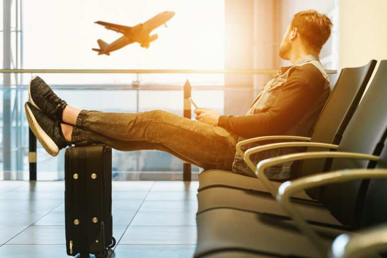 Persona viendo un avión por la ventana del aeropuerto, con los pies apoyados en la maleta. 